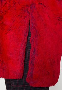 Tissu en fausse fourrure rouge avec une texture douce, présentant un ourlet fendu et un pantalon à carreaux noirs contrastants avec des accents roses.