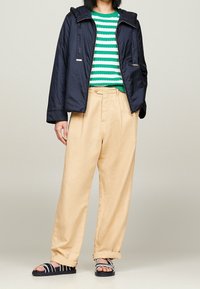 Personne portant une veste navy, un pull rayé vert et blanc, un pantalon large beige et des sandales noires avec des brides blanches, debout.