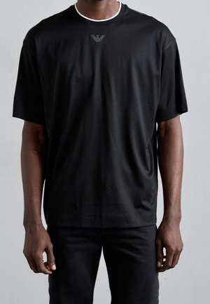 Homme portant un t-shirt noir à manches courtes avec un petit logo sous l'encolure et un pantalon noir devant un fond gris uni.