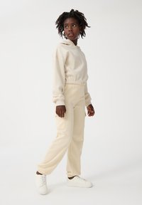 Beżowa bluza z kapturem o luźnym kroju i elastycznym pasku w talii, zestawiona z pasującymi beżowymi spodniami dresowymi z kieszeniami cargo, noszona z białymi trampkami.