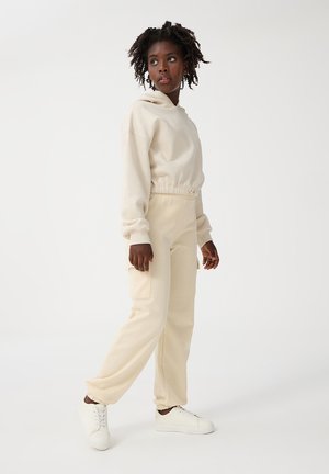Felpa beige con vestibilità comoda e vita elastica, abbinata a pantaloni della tuta beige con tasche cargo, indossata con sneakers bianche.