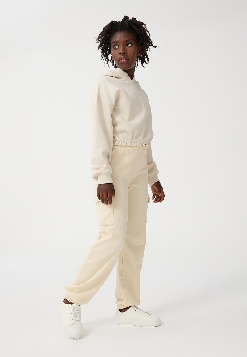 Beżowa bluza z kapturem o luźnym kroju i elastycznym pasku w talii, zestawiona z pasującymi beżowymi spodniami dresowymi z kieszeniami cargo, noszona z białymi trampkami.