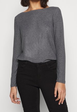 Vrouw die een grijze trui met lange mouwen draagt, ingestopt in een zwarte broek met hoge taille, gefotografeerd van nek tot heupen.