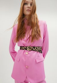 Rosa blazer med två framfickor, guldknappar och ett svart leopardmönstrat bälte med en guldlogobälte, stylad med lösa hår.