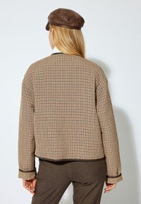 Quiltad långärmad jacka i ett brunt, krämfärgat och svart rutmönster. Har rund halsringning och elastiska ärmslut. En brun keps fulländar looken.