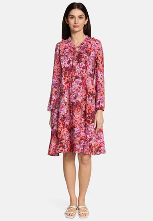 Woman wearing knee-length, long-sleeve floral dress in shades of red, pink, and purple, paired with white strappy sandals, standing.