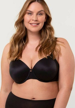 Kobieta o długich falowanych włosach uśmiechająca się, ubrana w czarny biustonosz na fiszbinach oraz pasujące wysokie majtki, na neutralnym jasnym tle.