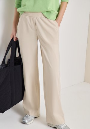 Person trägt beige Hosen mit weitem Bein, silberne Turnschuhe und grünen Pullover und hält eine große schwarze Tragetasche vor einem einfarbigen Hintergrund.