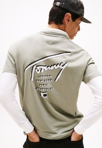 Olivgrünes Polohemd mit einer weißen Langarmunterlage; verfügt über einen großen schwarzen Grafischschriftzug auf dem Rücken mit Städtenamen und einem Logo.