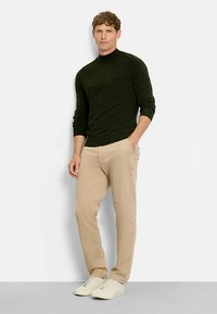 Dunkelgrüner Pullover mit langen Ärmeln und hohem Kragen, kombiniert mit hellbeigen Hosen und weißen Turnschuhen. Einfaches Design, glatte Textur.