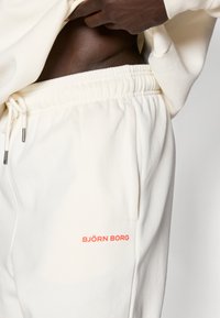 Κρεμ ζακέτες από μαλακό ύφασμα, με ελαστική μέση, κορδόνι, πλαϊνές τσέπες και αντίθετο πορτοκαλί λογότυπο.