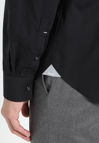 Svart langermet skjorte med knappede ermer, slim fit, og en grå etikett i sømmen, kombinert med grå bukser.