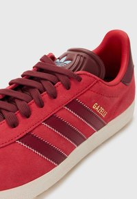 Red suede sneaker with burgundy accents, white rubber sole, textured laces, and embroidered "Gazelle" logo in gold. Classic three stripes design.