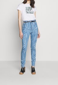 Jeans ajustados en denim azul claro con rayas verticales de cebra, combinados con una camiseta blanca que presenta un texto gráfico en verde, y sandalias negras de tacón.