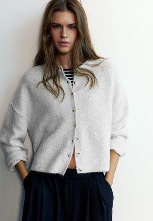 Jeune femme aux longs cheveux portant un cardigan gris clair boutonné par-dessus un top rayé et un pantalon foncé, les mains dans les poches.