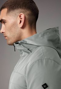 Jachetă impermeabilă de culoare gri deschis cu glugă, având închidere cu fermoar, un design elegant și un mic emblemă neagră pe umăr.