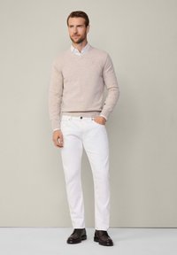Jasnobeżowy sweter dzianinowy na białej koszuli z kołnierzykiem, w połączeniu z białymi spodniami i ciemnobrązowymi butami. Prosty design, bez widocznych wzorów.