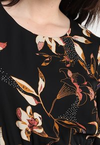 Blusa nera con motivo floreale, caratterizzata da grandi fiori e foglie beige, rosa e marroni, indossata da una persona con capelli scuri.