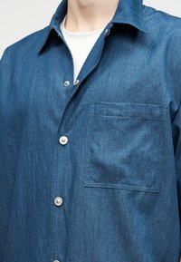 Κοντινό πλάνο ενός ατόμου που φοράει μπλε τζιν πουκάμισο με κουμπιά πάνω από λευκό μπλουζάκι με λαιμόκοψη.