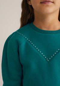 Felpa color teal con scollo a giro, caratterizzata da un tessuto testurizzato, maniche a sbuffo e una fila di borchie argentate lungo la cucitura della spalla.