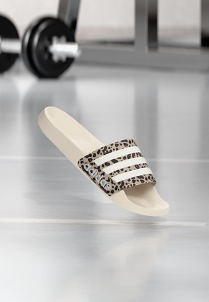 Sandale slide beige Adidas avec bride imprimé léopard et bandes blanches flottant au-dessus d’un sol brillant, poids de gym flous en arrière-plan.