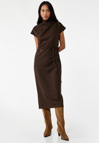 Vestido midi marrón con un escote alto, mangas cortas y un cinturón atado en la cintura; modelo lleva botas de rodilla beige.