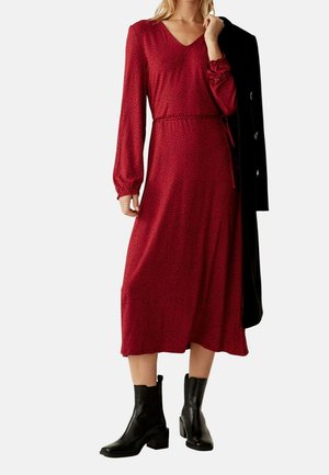 Kvinde iført en langærmet rød midi-kjole med sorte polkaprikker, sorte ankelstøvler og med en sort frakke over den ene skulder.
