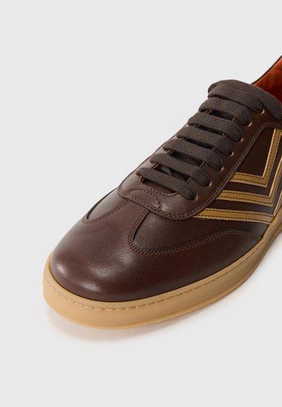Tenisi din piele maro cu talpă de cauciuc bej, având vârf rotund, accente texturate și șireturi maro. Model în zigzag de culoare bej deschis pe lateral.