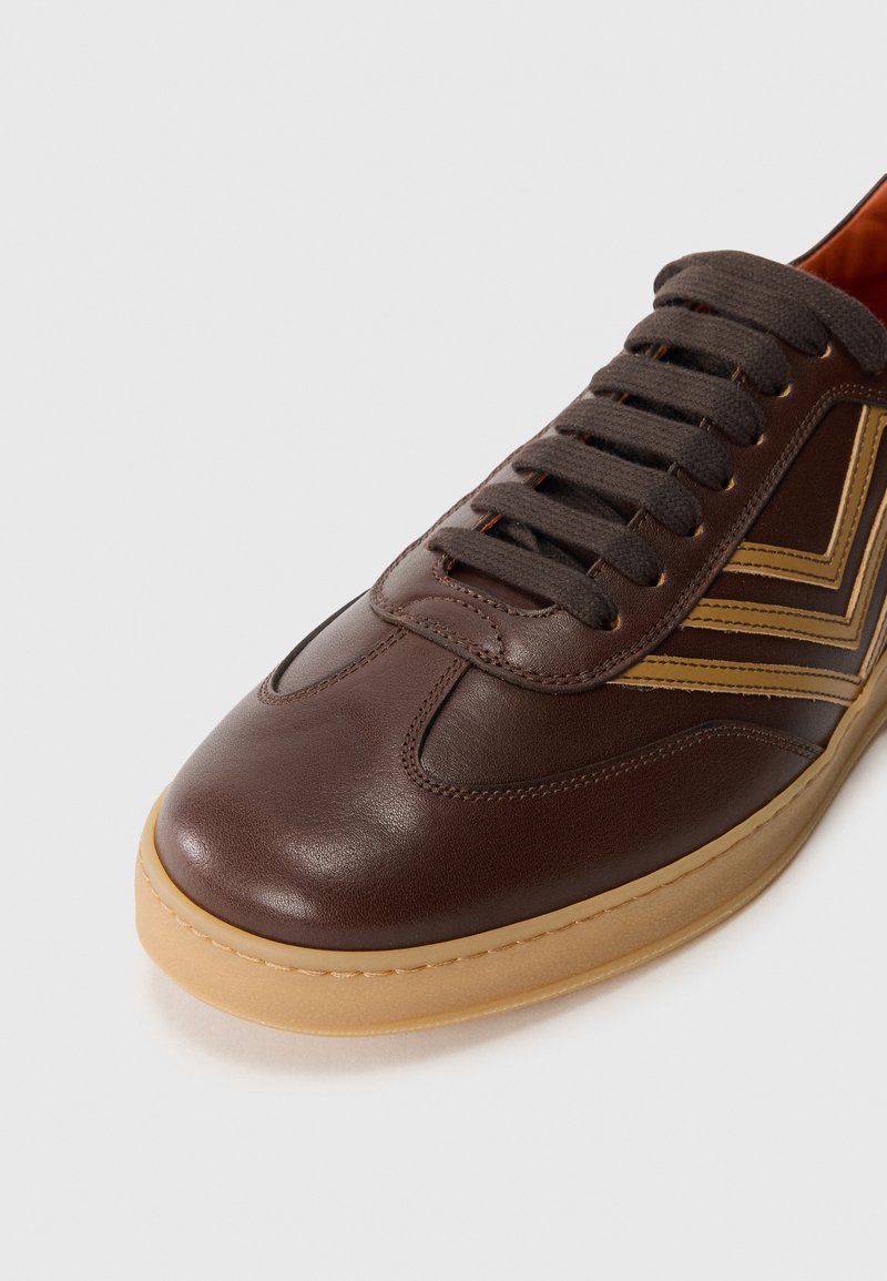 Sneaker in pelle marrone con suola in gomma beige, presenta punta rotonda, dettagli testurizzati e lacci marroni. Motivo a chevron in marrone chiaro sul lato.
