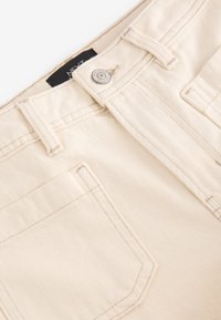 Pantalon en coton beige avec une finition texturée, comprenant un bouton en métal, des passants de ceinture et des poches avant. Les coutures mettent en valeur les détails du design.