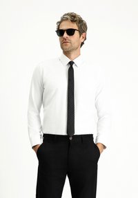 Camicia bianca a maniche lunghe abbinata a una cravatta nera a quadretti e pantaloni neri. Modello con occhiali da sole neri, mani nelle tasche.
