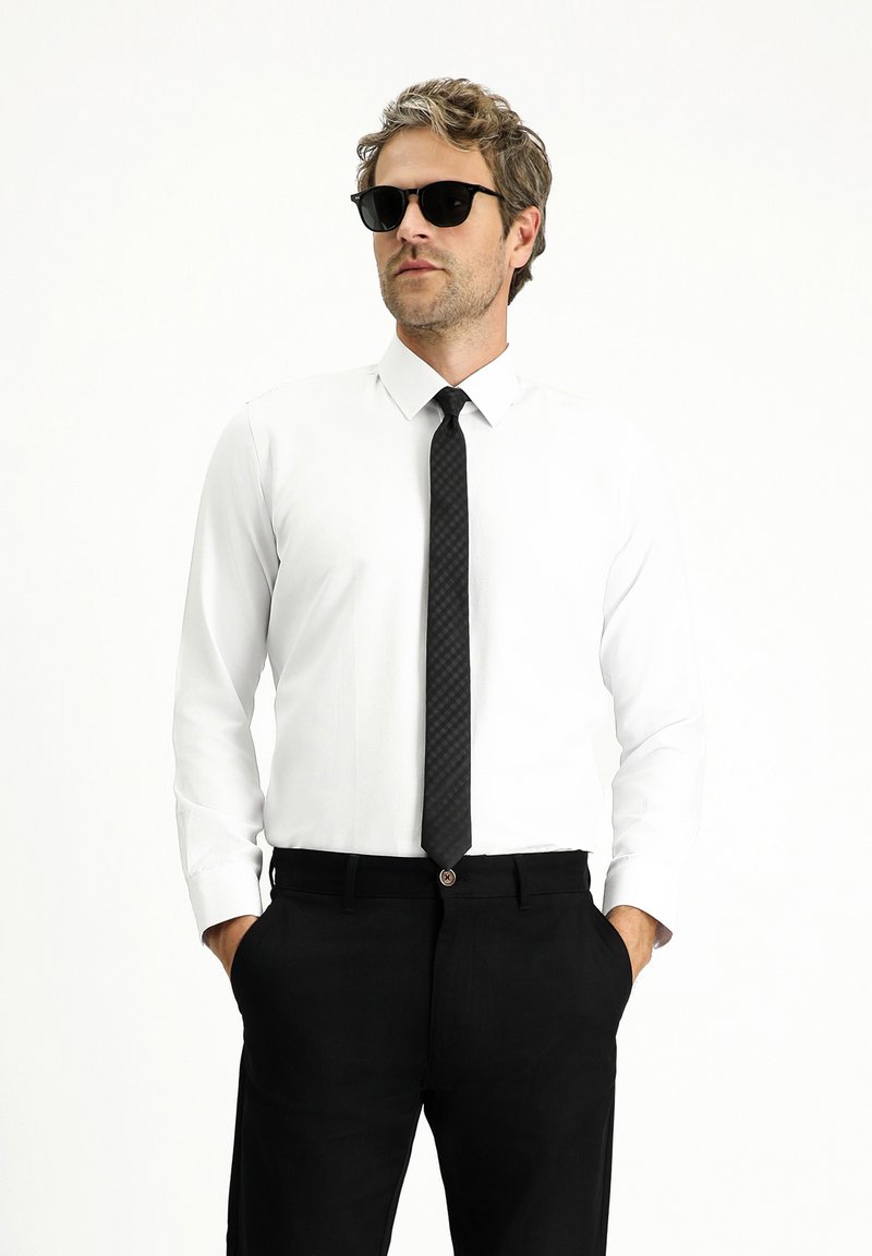 Camicia bianca a maniche lunghe abbinata a una cravatta nera a quadretti e pantaloni neri. Modello con occhiali da sole neri, mani nelle tasche.