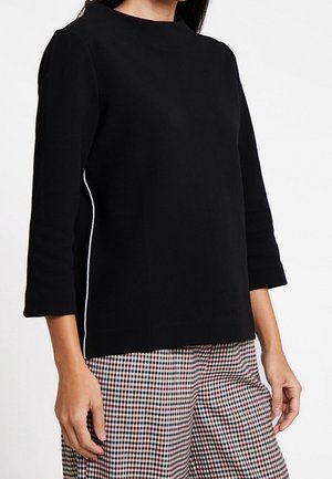 Vrouw draagt een zwart shirt met lange mouwen met witte bies aan de zijkant en geruite broek met zwart-rood-witte patronen.