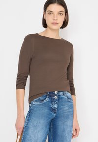 Hnědá košile s dlouhými rukávy, kulatým výstřihem a vyhrnutými rukávy, kombinovaná se světle modrými džínami s dvěma předními knoflíky a detaily kapes.