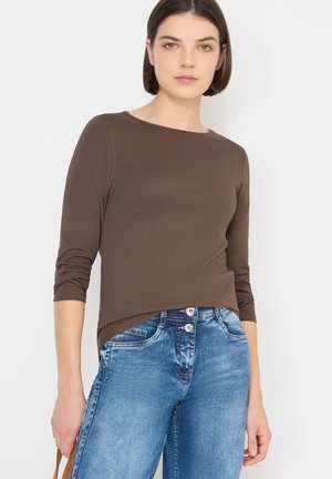 Hnědá košile s dlouhými rukávy, kulatým výstřihem a vyhrnutými rukávy, kombinovaná se světle modrými džínami s dvěma předními knoflíky a detaily kapes.