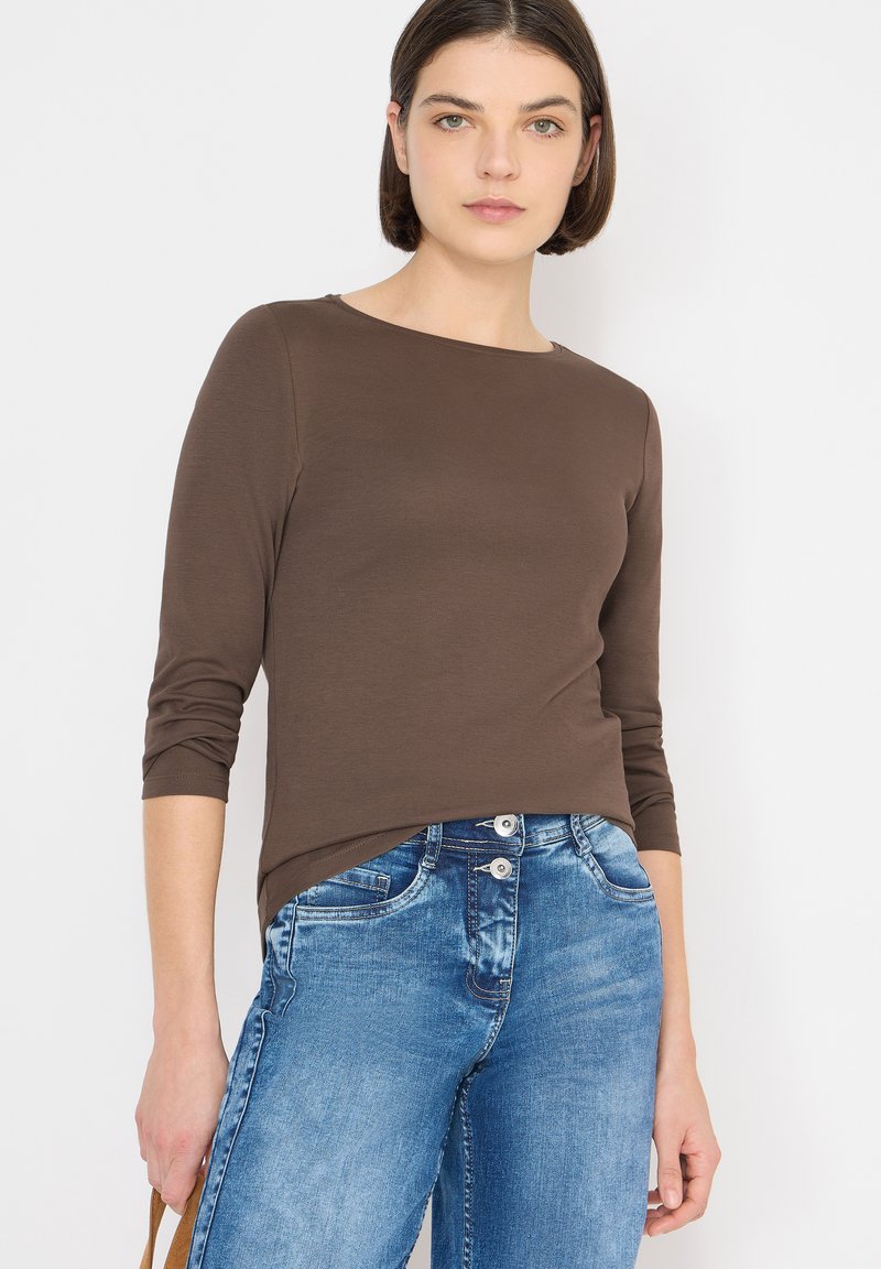 Hnědá košile s dlouhými rukávy, kulatým výstřihem a vyhrnutými rukávy, kombinovaná se světle modrými džínami s dvěma předními knoflíky a detaily kapes.