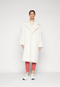 Cappotto lungo bianco e fluffy con ampio colletto, chiusura a bottoni, tasche laterali, indossato sopra una maglia chiara e leggings rosa, abbinato a sneaker bianche.