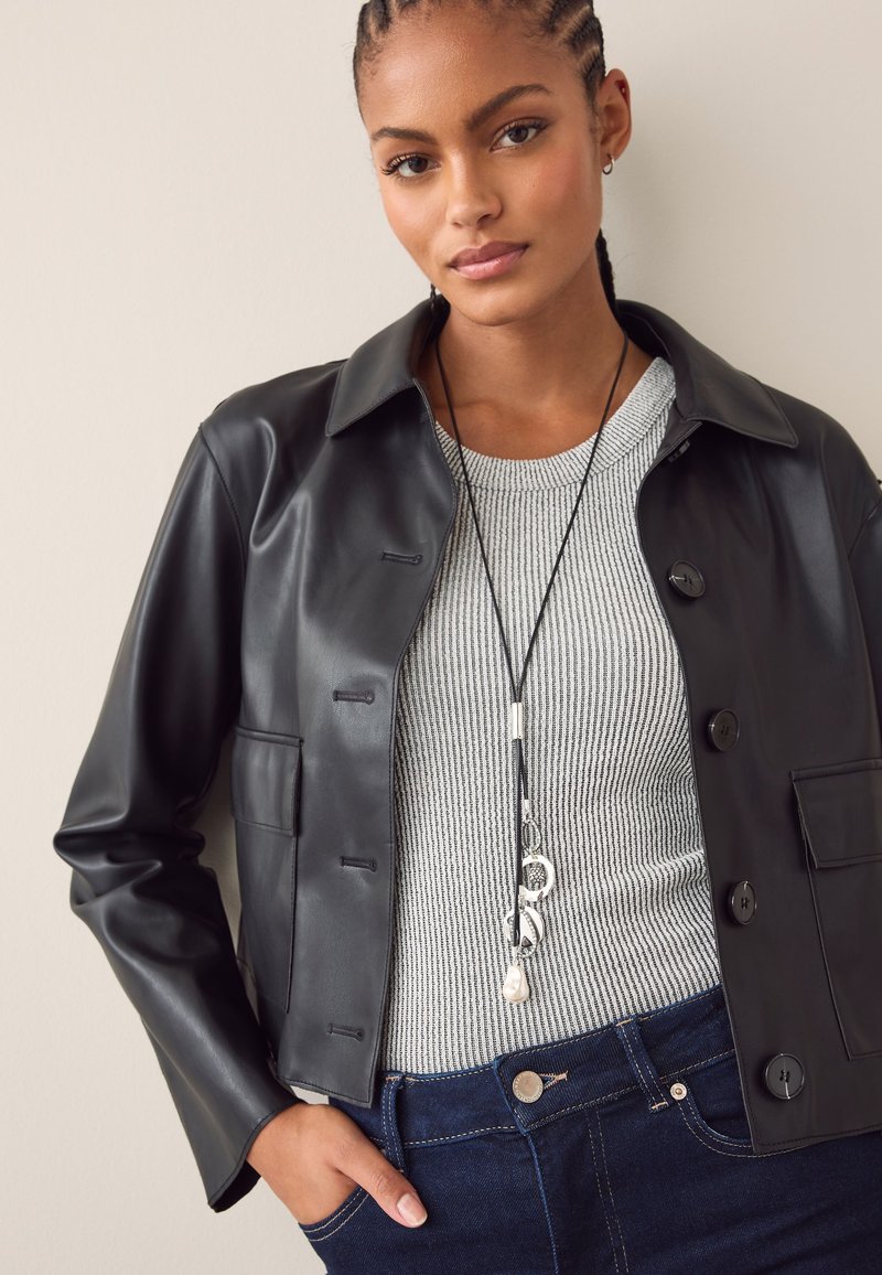 Schwarze Jacke aus Kunstleder mit Kragen, ausgestattet mit Knopfverschluss und einer Brusttasche, kombiniert mit einem grauen, gerippten Pullover und dunklen Jeans.