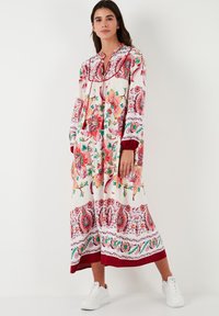 Vestido maxi floral con base blanca, flores rojas y rosas, y bordes con patrones intricados. Presenta un collar y mangas largas, combinado con zapatillas blancas.