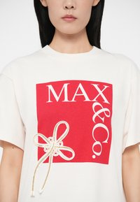 Λευκό T-shirt με μεγάλο κόκκινο τετράγωνο που έχει το κείμενο "MAX & Co." και διακοσμητική χρυσή κορδέλα στην αριστερή πλευρά, φορεμένο από ένα άτομο.