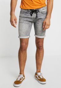 Pantalones cortos de mezclilla gris con dobladillos enrollados, con una cintura ajustable mediante cordón. Combinados con zapatillas blancas y una camiseta naranja.