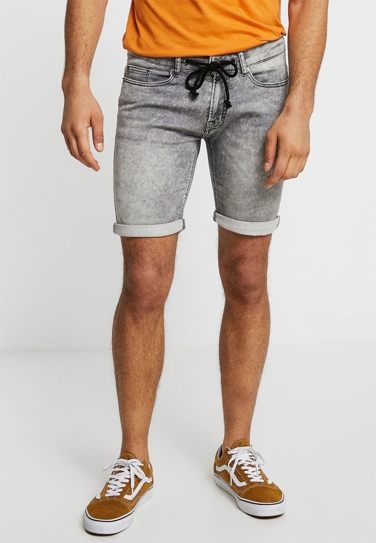 Pantalones cortos de mezclilla gris con dobladillos enrollados, con una cintura ajustable mediante cordón. Combinados con zapatillas blancas y una camiseta naranja.