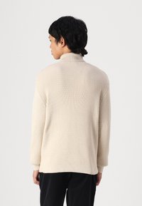 Sweater de malha bege com um colarinho alto, apresentando um padrão texturizado, corte descontraído e punhos canelados, visto de costas.