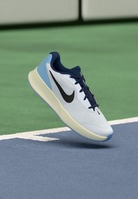 Adidași pentru alergare cu sireturi de culoare bleumarin, parte superioară din plasă ușoară albastră și albă, și logo-ul Nike în negru, pe un teren sportiv albastru și verde.