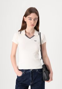 Kremasta polo majica s tamnoplavim detaljima i rebrastom teksturom, u kombinaciji s tamnoplavim trapericama. Model drži malu crnu torbu.
