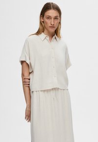 Light beige short-sleeve shirt made of a textured fabric with a button-down front and a cropped hem. Paired with a matching light beige skirt.