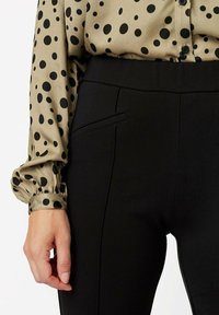 Beżowa bluzka w czarne kropki z bufkami, zestawiona z dopasowanymi czarnymi spodniami z boczną kieszenią i gładką fakturą.