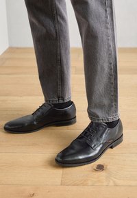 Chaussures habillées en cuir noir avec un bout pointu, dotées d'un design à lacets et d'un marquage discret sur le côté. Portées avec un pantalon en denim gris.