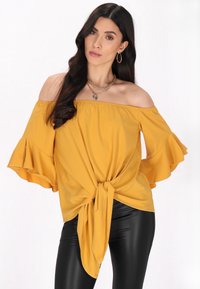 Gele off-the-shoulder top met volumineuze mouwen en een geknoopte voorkant, gemaakt van lichtgewicht stof. Gecombineerd met zwarte leren leggings.