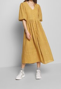Senapsgul midi-klänning med v-ringning, puffärmar och knapprad. Har en strukturerad, perforerad design som kombineras med vita sneakers.